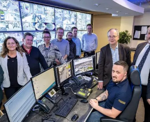 Councillor David Neighbour, Leader of Hart District Council, and Councillor Stuart Bailey, portfolio holder for community at Hart District Council, at the Runnymede CCTV control centre with a bank of monitors and group of councillors and council officers