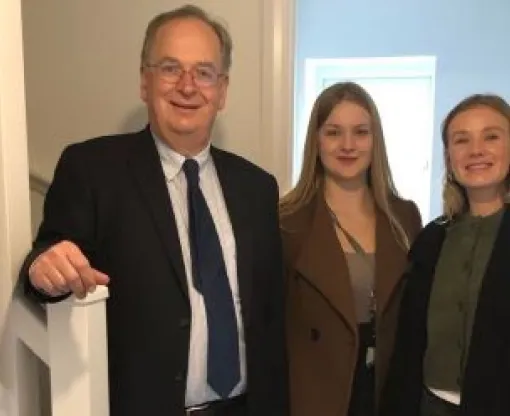 Man standing in the hallway of a house with his right hand on the stairs bannister with two women beside him