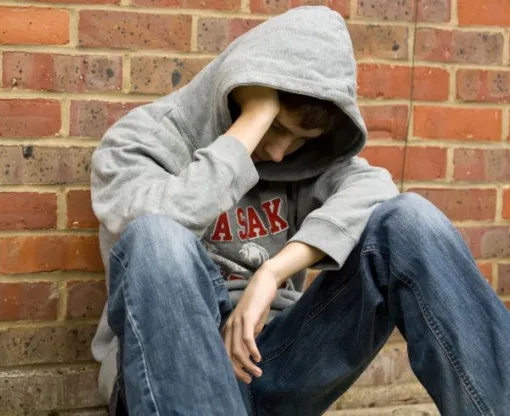 Boy wearing grey sweatshirt and jean with hood pulled over face sitting against a red brick wall