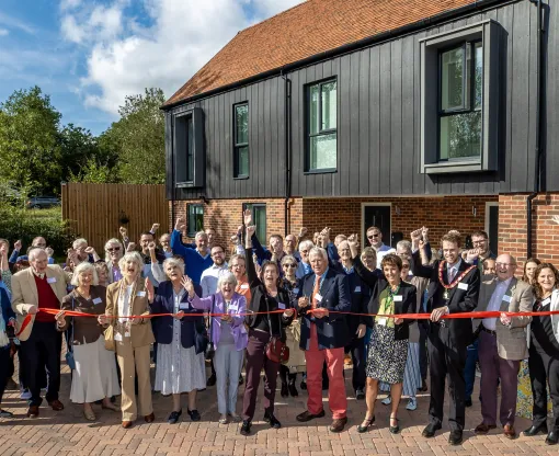 Group of people outside a brick and wood holding a red ribbon which a man is cutting with scissors