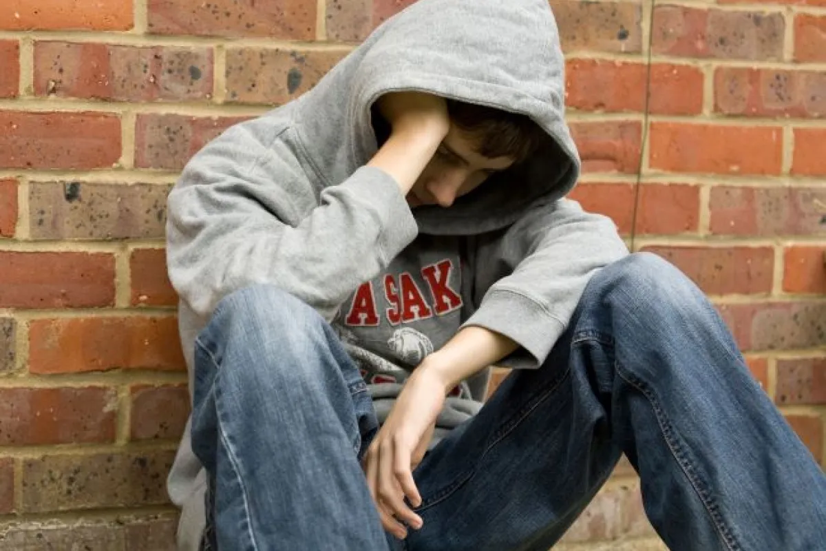 Boy wearing grey sweatshirt and jean with hood pulled over face sitting against a red brick wall