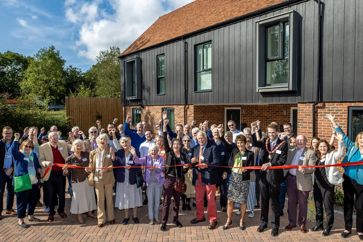 Group of people outside a brick and wood holding a red ribbon which a man is cutting with scissors