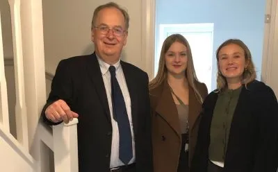 Man standing in the hallway of a house with his right hand on the stairs bannister with two women beside him