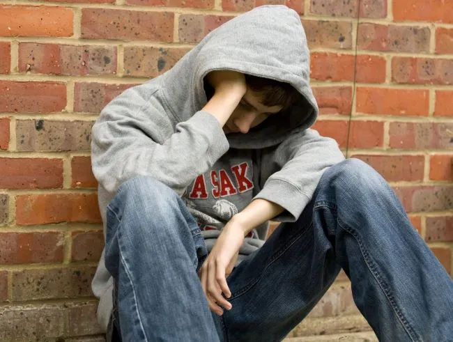 Boy wearing grey sweatshirt and jean with hood pulled over face sitting against a red brick wall