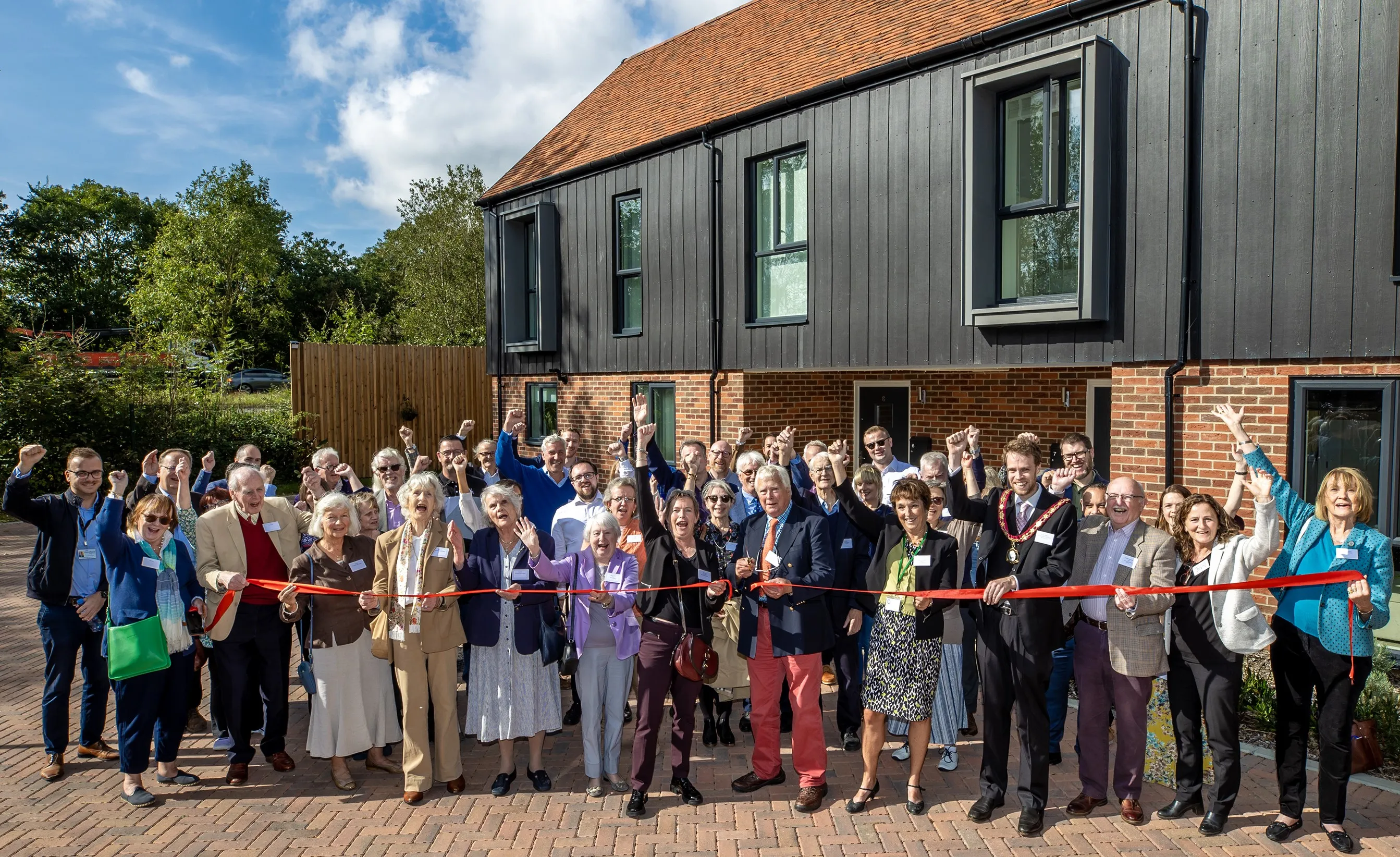 Group of people outside a brick and wood holding a red ribbon which a man is cutting with scissors