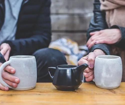 Two people having a hot drink with hands around two mugs and a jug